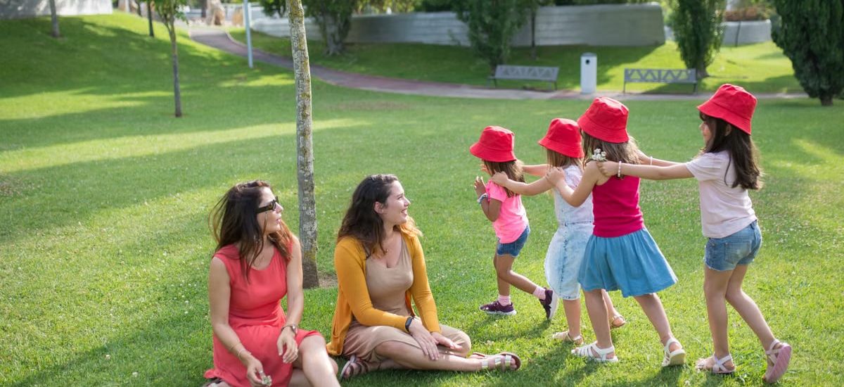 Happy children playing in a sunny park with two mothers sitting nearby, enjoying the outdoors.